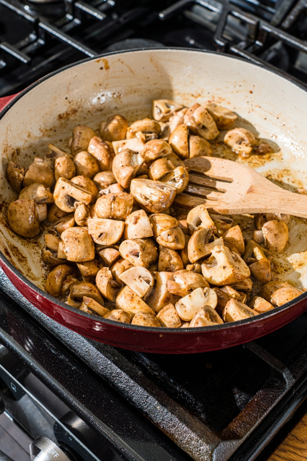 A cast iron skillet with a wooden spoon stirring mushrooms. The skillet is on a stove.