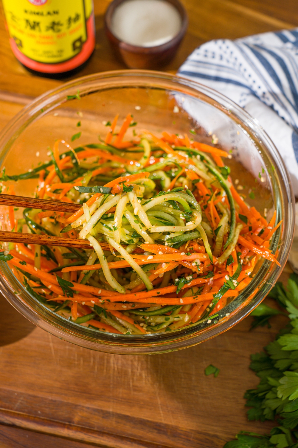A glass bowl with shredded carrots and cucumber with a pair of chopsticks tossing the vegetables in dressing. The bowl is on a wooden cutting board.