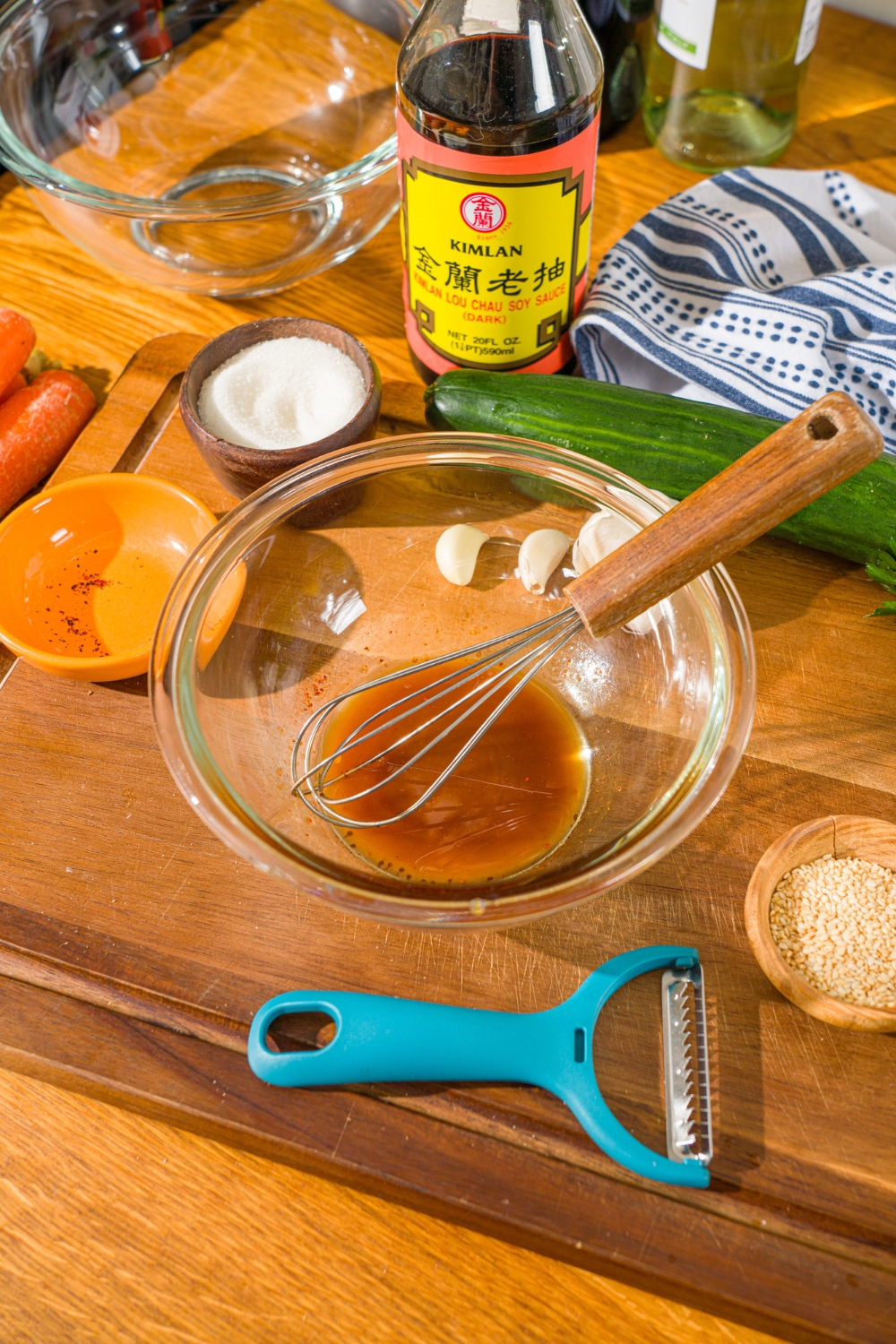 A glass bowl with a whisk mixing carrot cucumber salad dressing. The bowl is on a wooden counter iwth a cucumber and small bowls of ingredients.