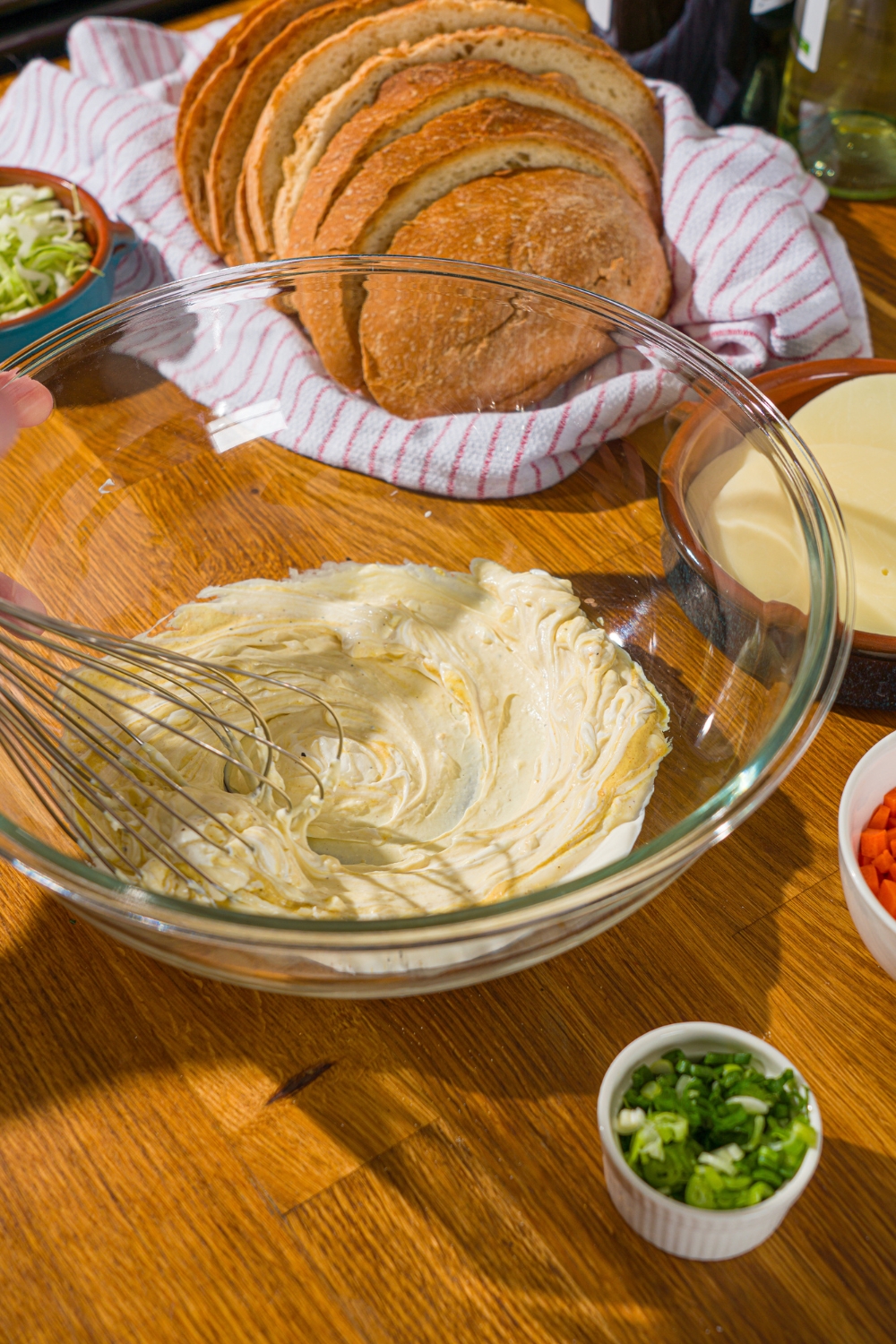 A glass bowl with a whisk mixing ingredients to make a dressing for a chicken melt sandwich. The bowl is on a wooden counter with a basket of bread and several bowls of ingredients.