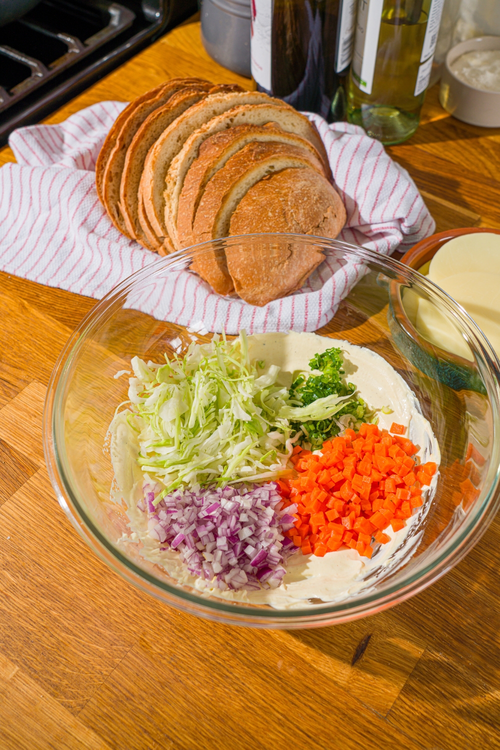 A glass bowl with chicken melt dressing topped with chopped vegetables like carrots, red onion, and cabbage. The bowl is on a wooden counter with a basket of sliced bread.