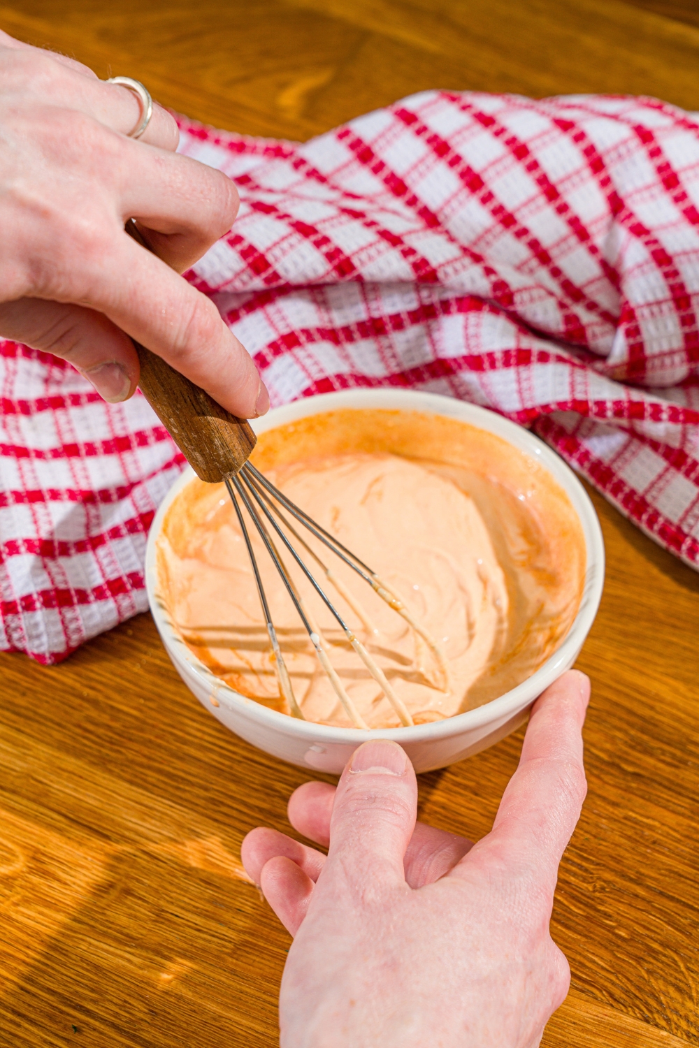 A white bowl with a whisk mixing ingredients to make comeback sauce. The bowl is on a wooden counter with a red checkered napkin.