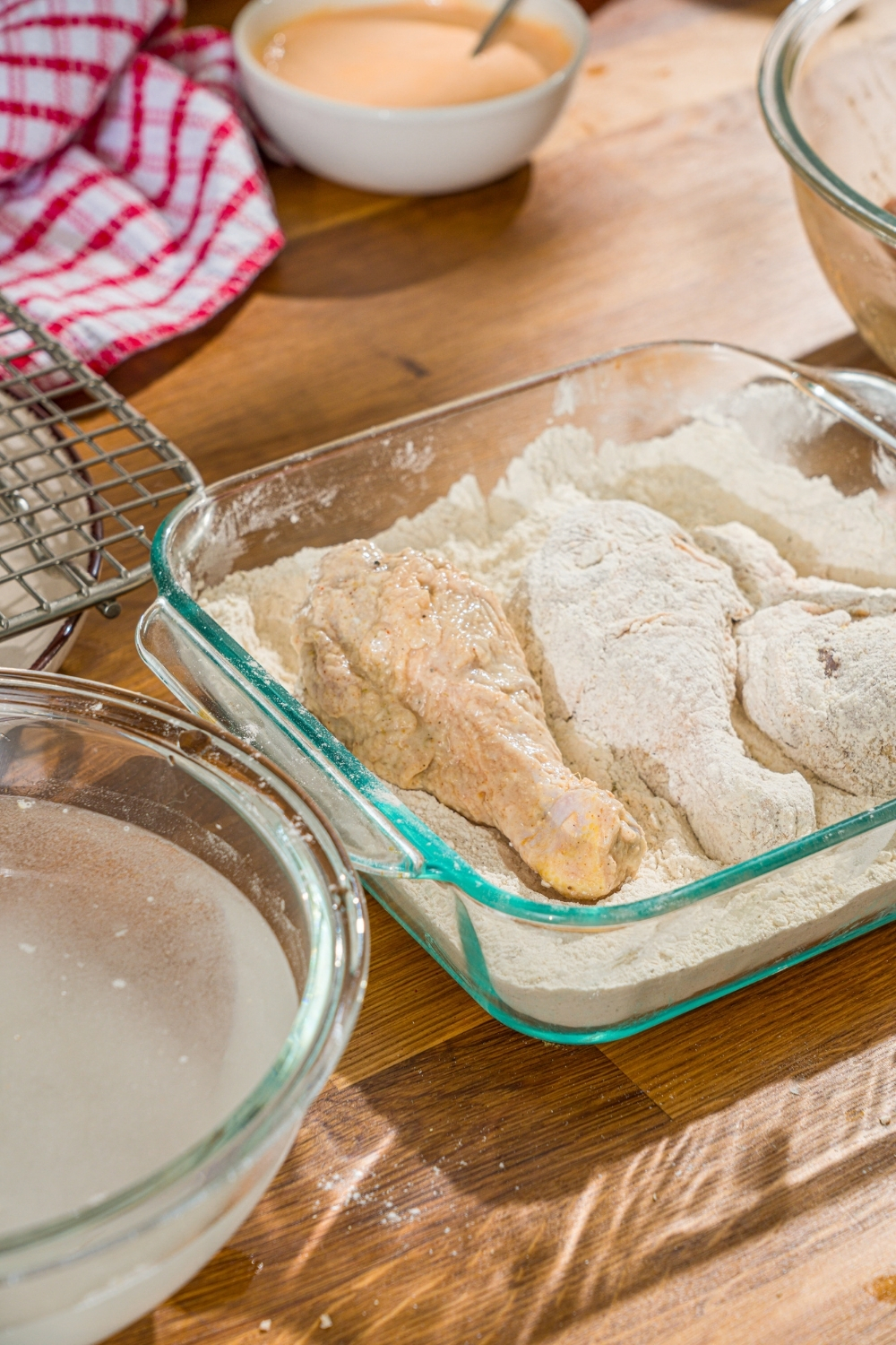 A glass baking dish with seasoned flour with several pieces of chicken. The dish is on a wooden counter with a bowl of water.