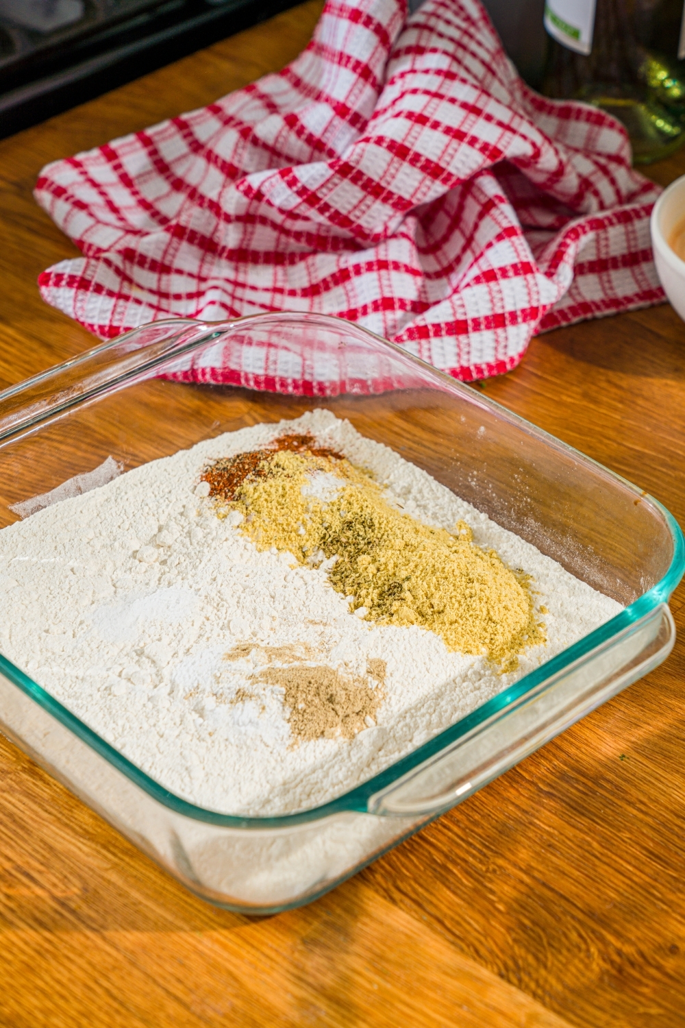 A glass baking dish with flour and seasonings including paprika, white pepper, garlic powder, chicken powder, oregano, and more. The dish is on a wooden counter with a red checkered napkin.