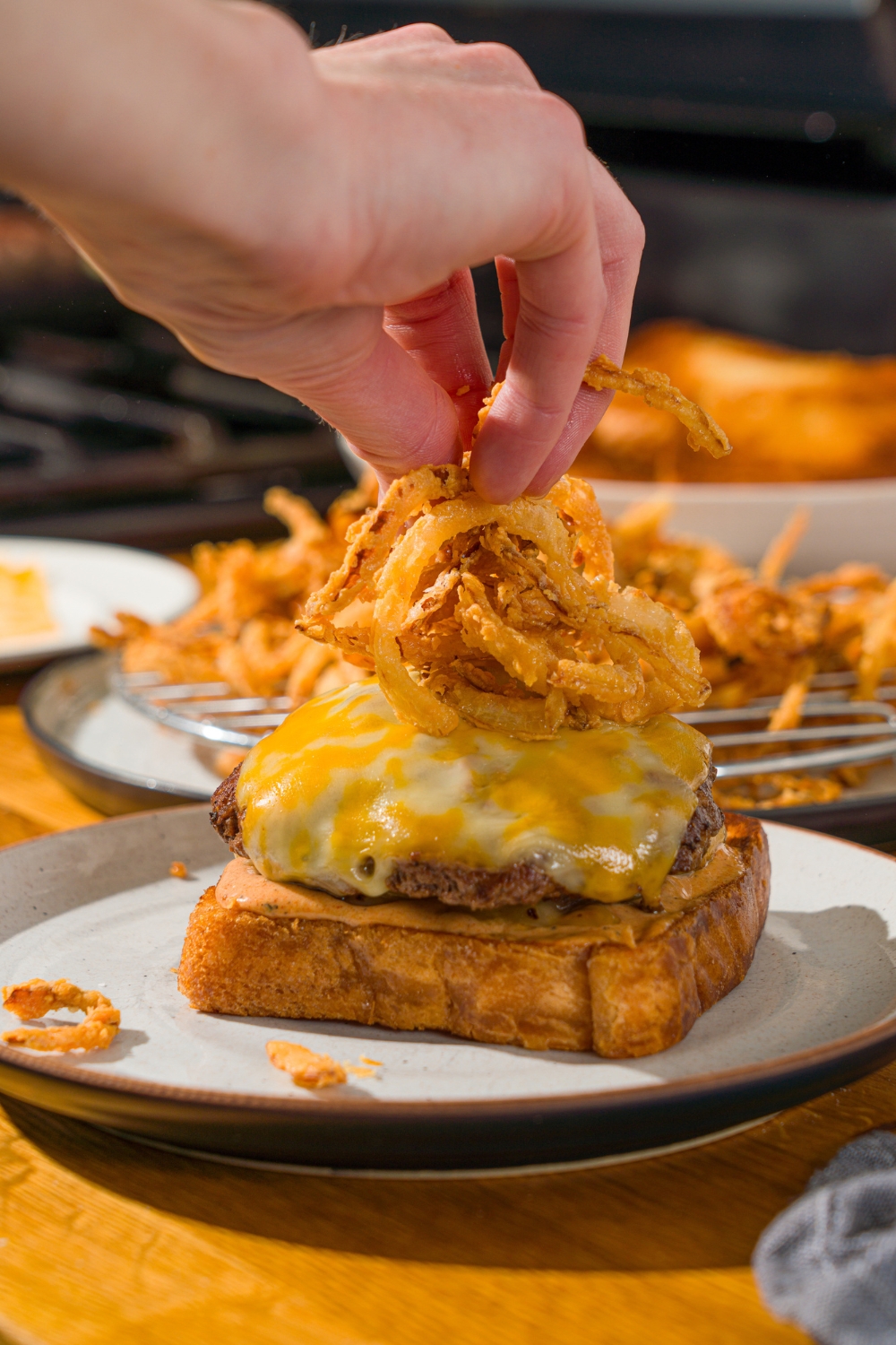 A plate with an open copycat Whataburger patty melt. A hand is adding fried onions to the top of the burger patty. The plate is on a wooden counter.