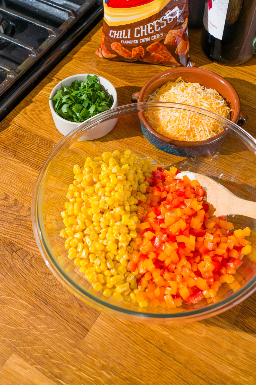 A glass bowl with a spoon mixing corn and diced red and yellow bell pepper. The bowl is on a wooden counter with bowls of ingredients.