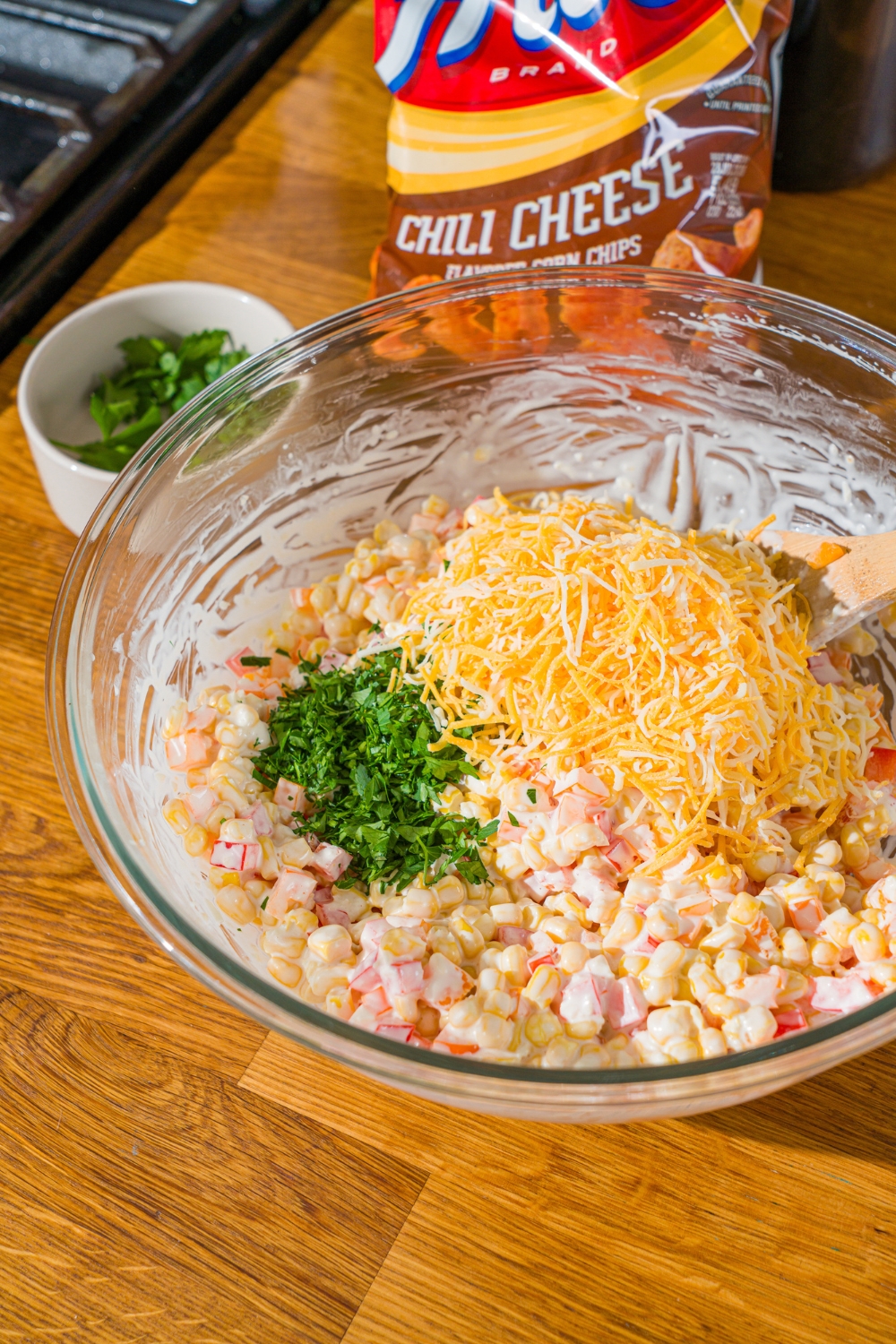 A glass bowl with corn and diced peppers tossed in a sour cream dressing. There is shredded cheese and chopped parsley on top. The bowl is on a wooden counter.