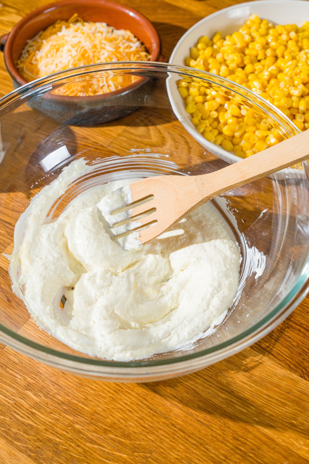 A glass bowl with a wooden spoon mixing corn salad sour cream dressing. The bowl is on a wooden counter with bowls of ingredients.