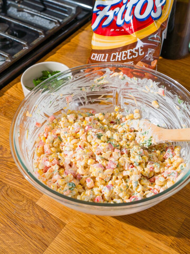 A glass bowl with a wooden spoon mixing corn, shredded cheese, and diced peppers in a sour cream dressing. The bowl is on a wooden counter with a bag of fritos.