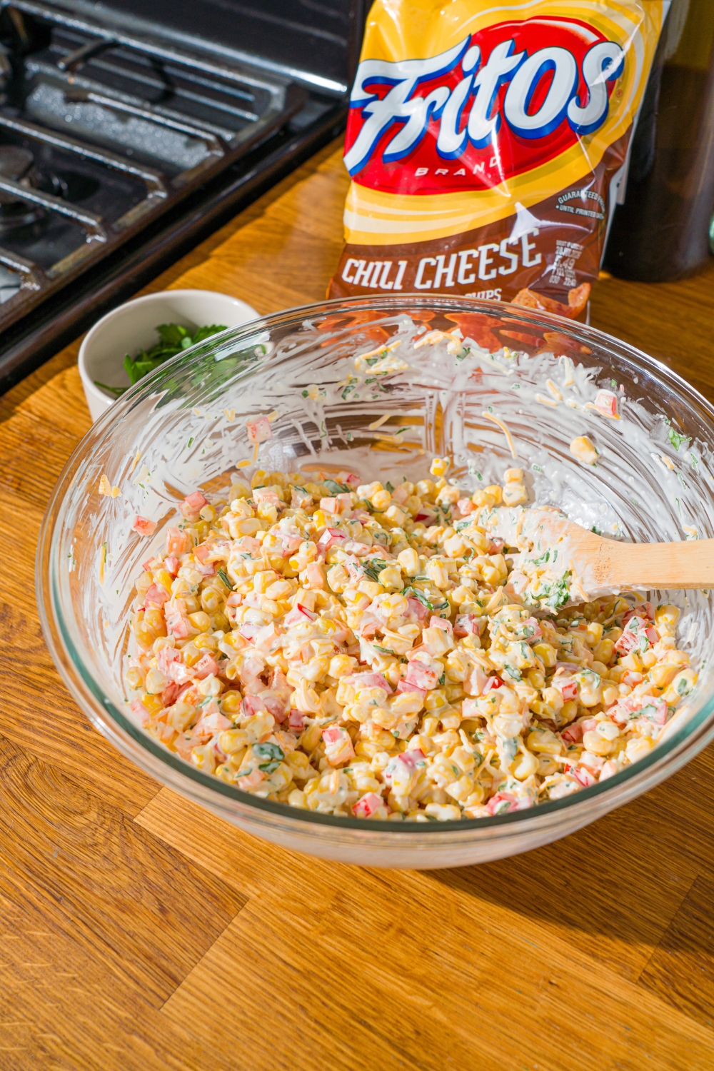 A glass bowl with a wooden spoon mixing corn, shredded cheese, and diced peppers in a sour cream dressing. The bowl is on a wooden counter with a bag of fritos.