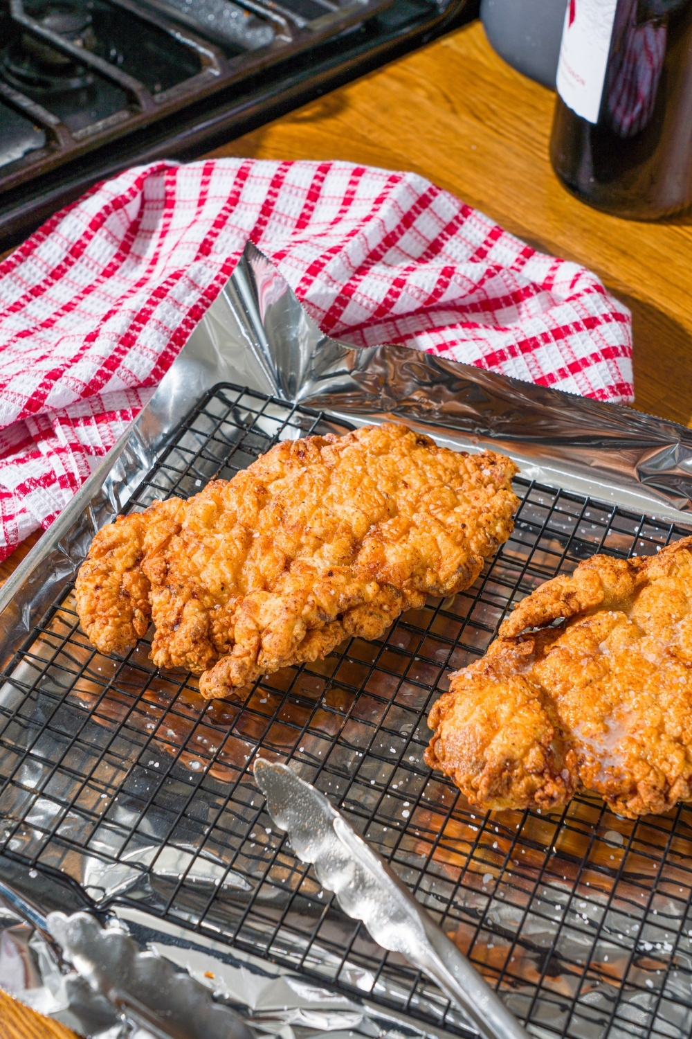 Two pieces of country fried chicken on a wire rack over a baking sheet and foil. The sheet is on a wooden counter with a red striped napkin.