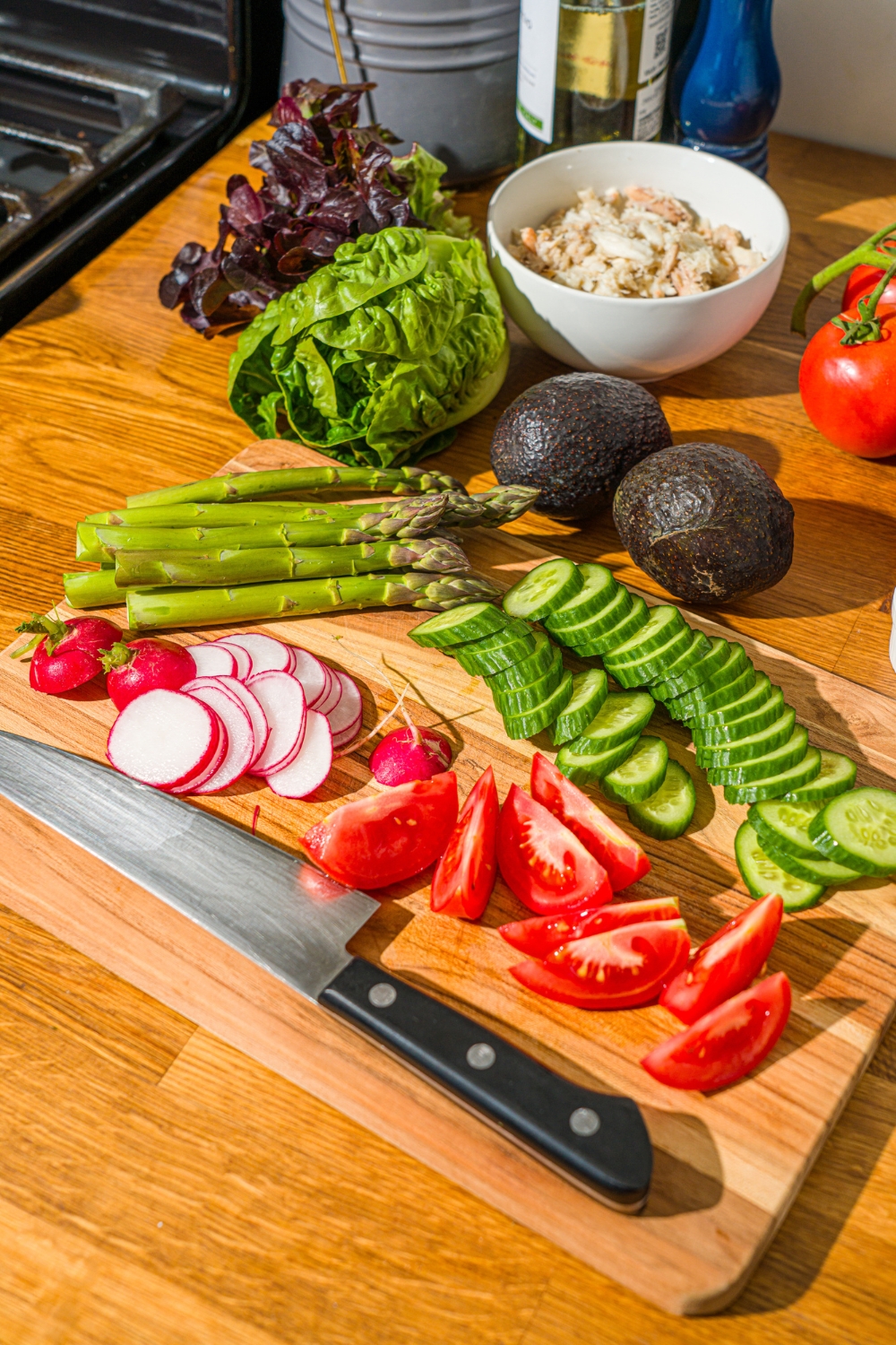 A wooden cutting board with a knife and sliced cucumber, tomatoes, and radishes, and asparagus. The board is on a wooden counter with a head of lettuce, avocados, and a bowl of lump crab.