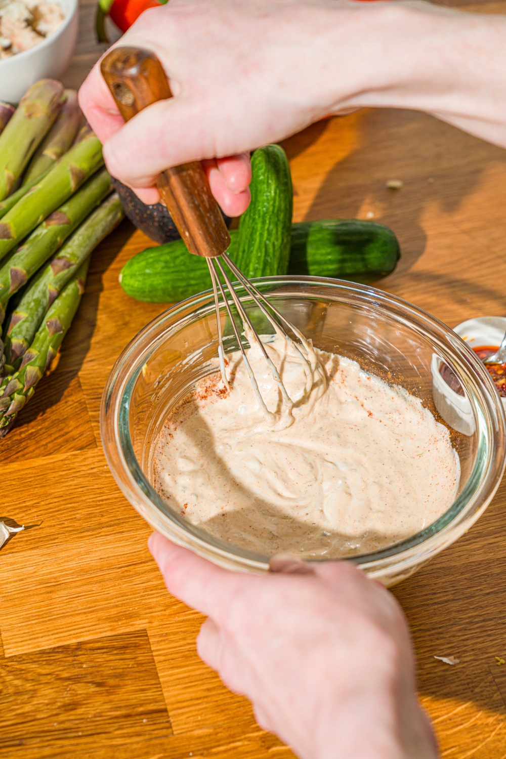 A glass bowl with a whisk mixing ingredients to make crab Louie salad dressing. The bowl is on a wooden counter with cucumbers and asparagus.
