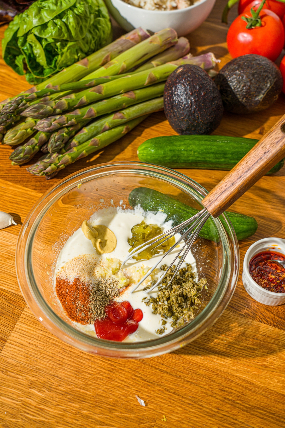 A glass bowl with a whisk and ingredients to make crab Louie salad dressing including mayo, greek yogurt, relish, mustard, ketchup, and seasonings. The bowl is on a wooden counter with cucumbers, asparagus, avocados, and more.