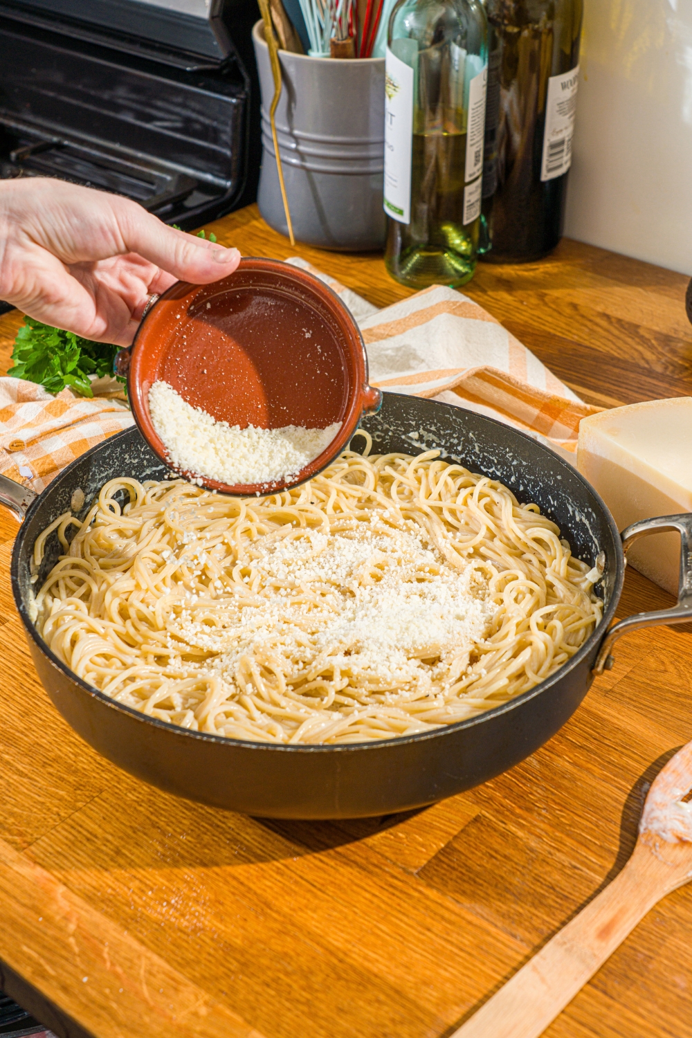 A skillet with cooked spaghetti in a creamy garlic sauce. A hand is pouring a bowl of parmesan cheese into the skillet. The skillet is on a wooden counter.