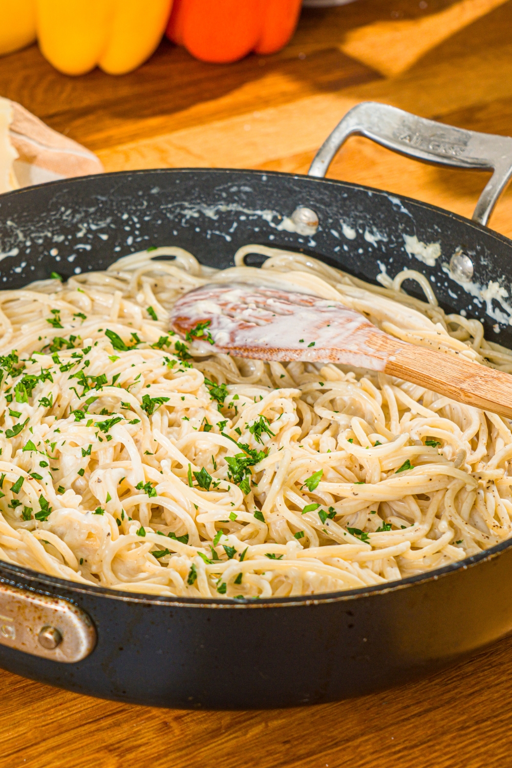 A skillet with spaghetti cooked in a creamy garlic parmesan sauce garnished with fresh parsley. A wooden spoon is stirring the mixture. The skillet is on a wooden counter.