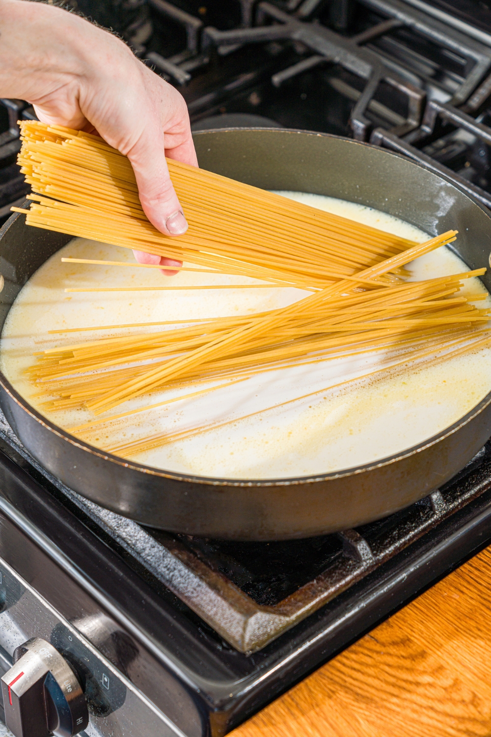 A skillet with a creamy garlic sauce base with uncooked pasta being added to the skillet. The skillet is on a stove.
