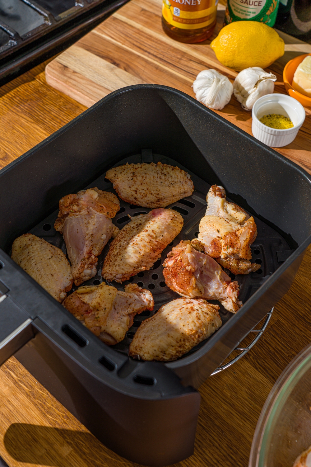 An air fryer tray with seasoned uncooked wings. The air fryer is on a wooden counter with a board of ingredients.
