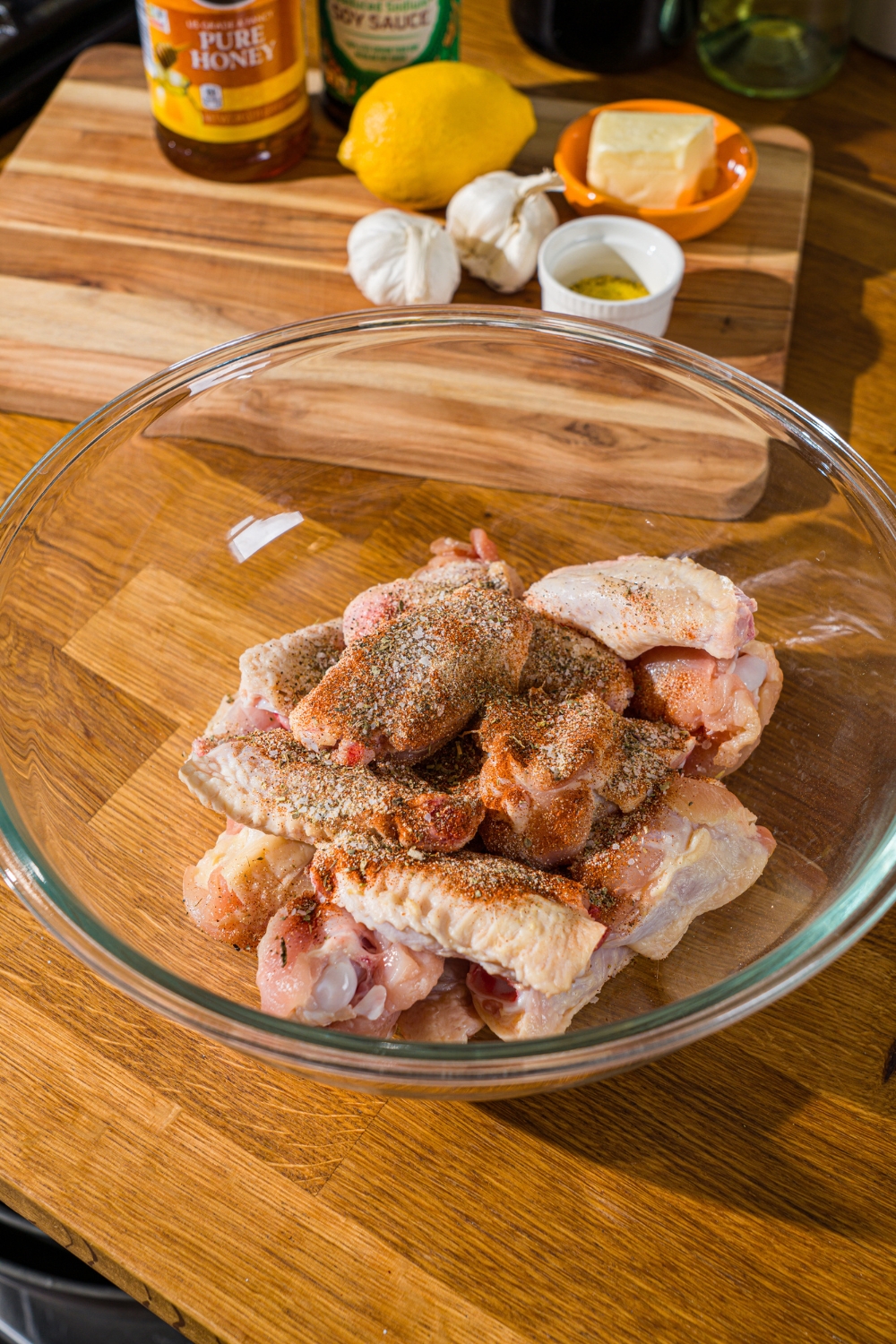 A glass bowl with uncooked chicken wings topped with a blend of seasonings. The bowl is on a wooden counter with a board of ingredients.