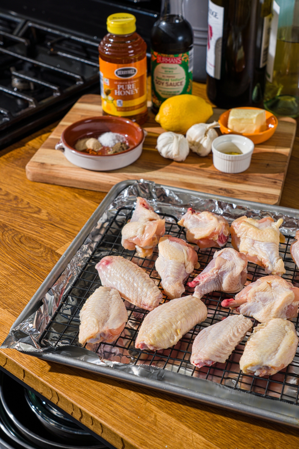 A baking sheet lined with foil with a tray of uncooked chicken wings on top. The tray is on a wooden counter with a board of ingredients.