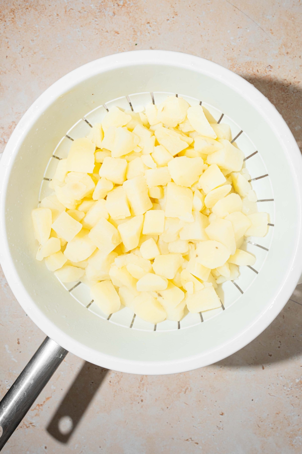 A strainer with cubed boiled potatoes. The strainer over a pot on a tan counter.