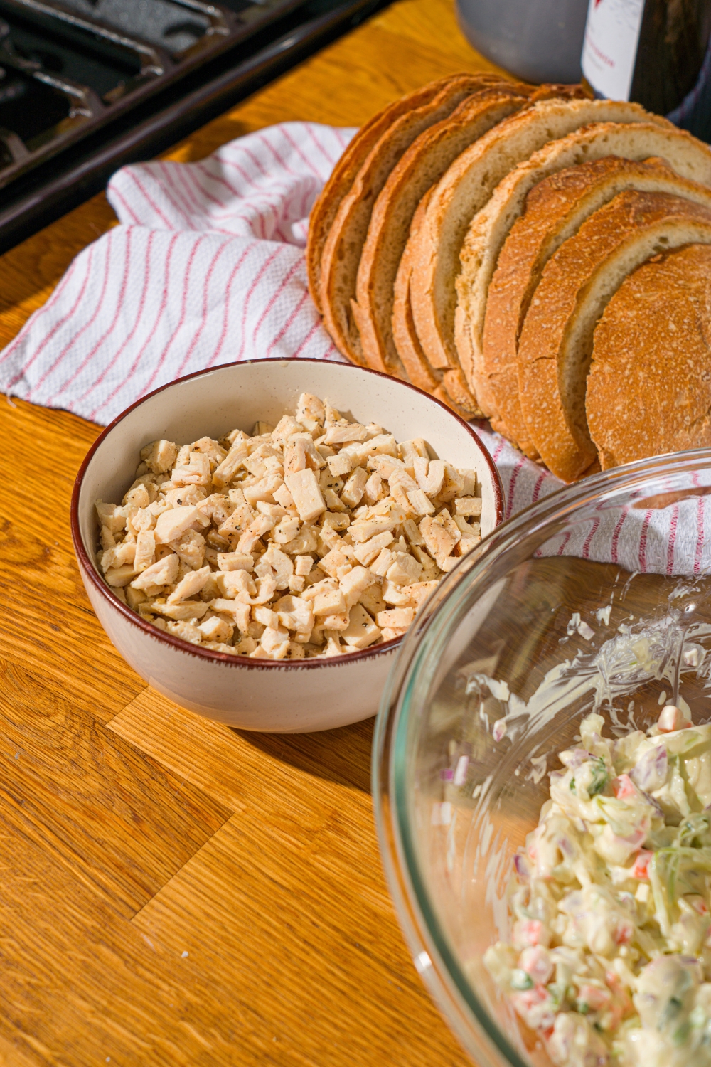 A white bowl of chopped cooked chicken. The bowl is on a wooden counter with a basket of sliced bread.