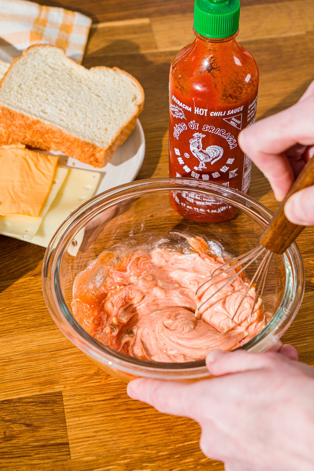 A glass bowl with a whisk mixing frisco sauce. The bowl is on a wooden counter with a jar of sriracha and plate of ingredients.