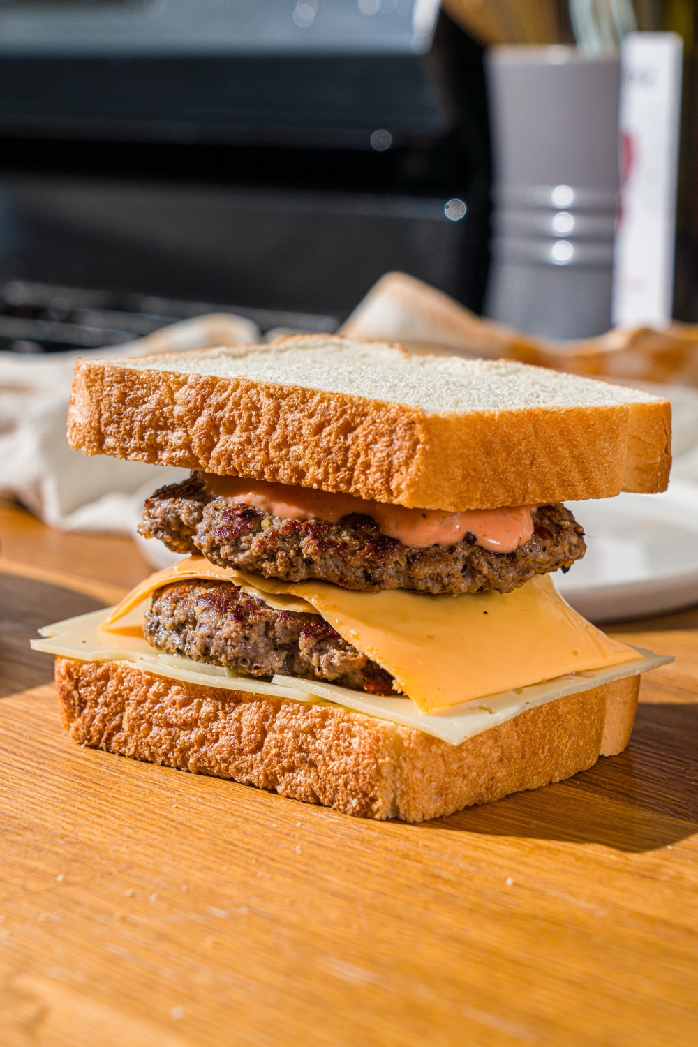 A wooden counter with an unmelted frisco melt including white bread, two burger patties, American cheese, swiss cheese, and frisco sauce.