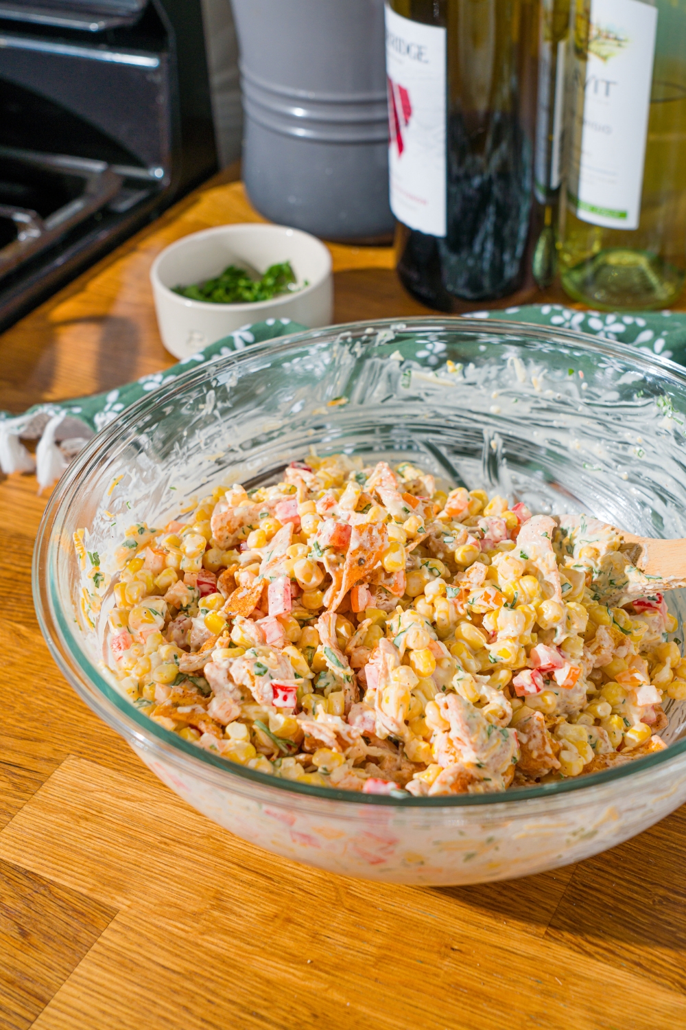 A glass bowl with a wooden spoon mixing corn, shredded cheese, diced peppers, and chili cheese fritos in a sour cream dressing. The bowl is on a wooden counter.