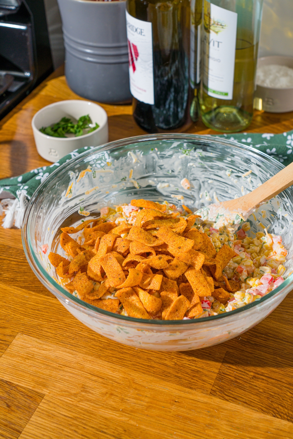 A glass bowl with a corn and pepper mixture tossed in sour cream dressing with chili cheese fritos on top. The bowl is on a wooden counter.