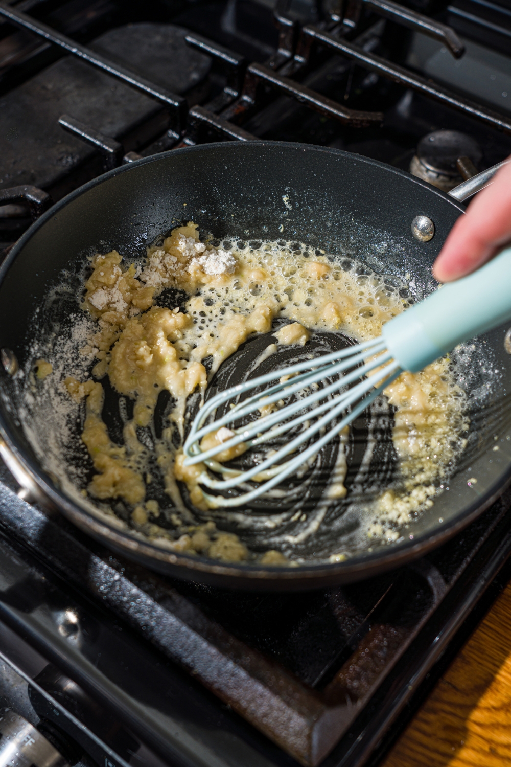 A skillet with a whisk mixing a garlic roux as it cooks on the stove.