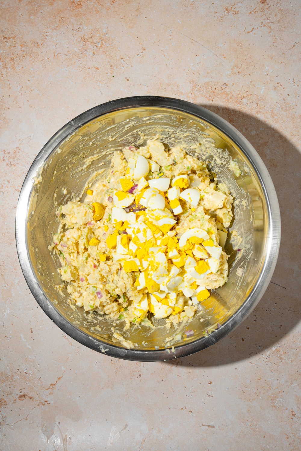 A large bowl with potato salad tossed in dressing with chopped egg on top. The bowl is on a tan counter.
