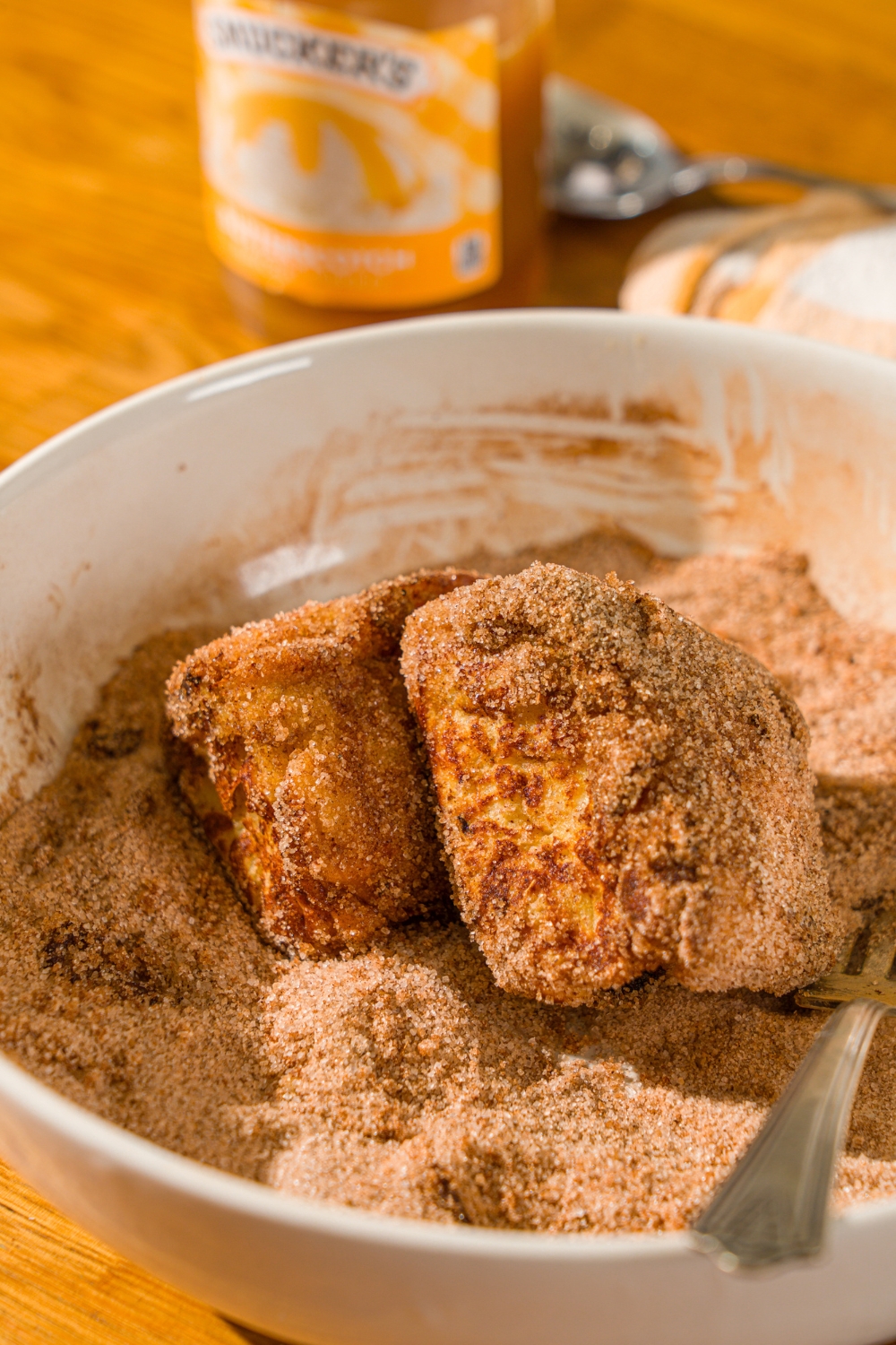 Two Hawaiian roll french toast bites in a large bowl of cinnamon sugar. The bites are coated with the mixture.