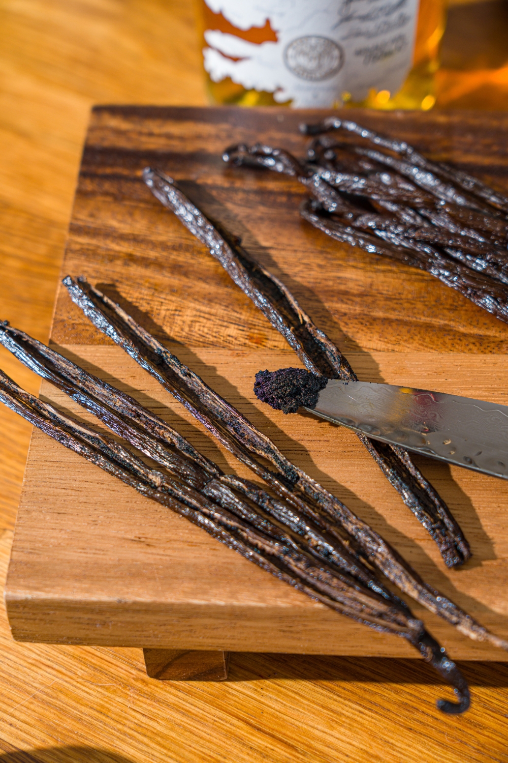 A wooden board with a knife paring vanilla bean pods and removing the seeds. The board is on a wooden counter.