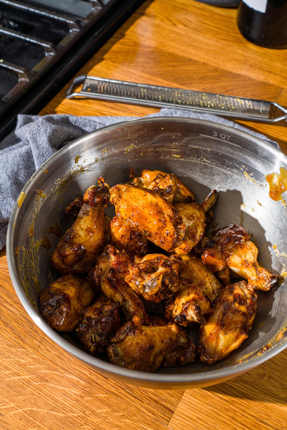 A large bowl with air fried chicken wings tossed in honey lemon pepper sauce. The bowl is on a wooden counter with a zester and blue cloth napkin.