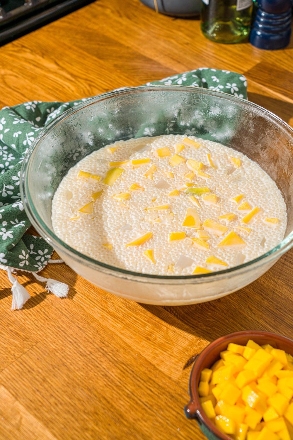 A glass bowl with mango sago with diced mango. The bowl is on a wooden counter with a green floral napkin and small bowl of diced mango.