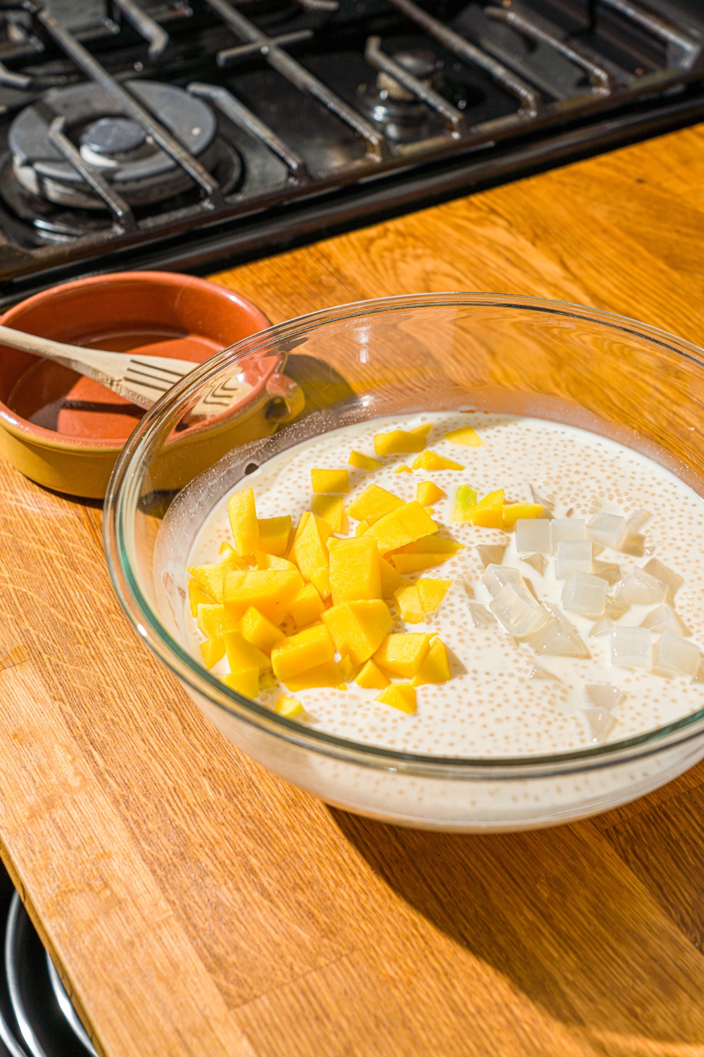 A glass bowl with a cooked sago mixture with diced mango and coconut jelly. The bowl is on a wooden counter.