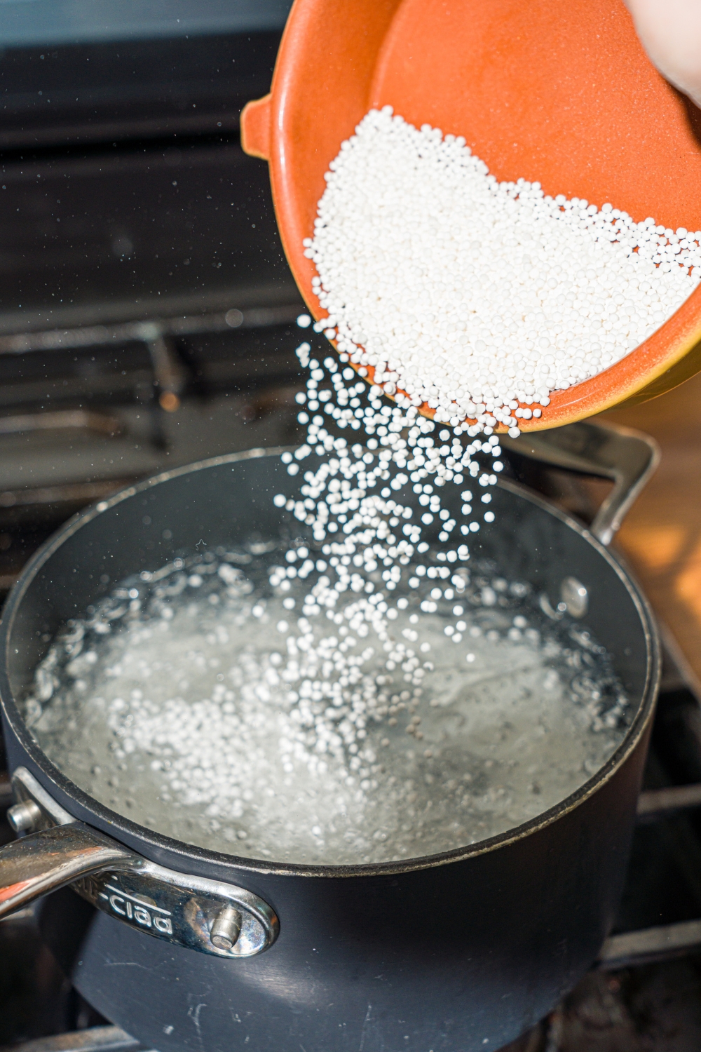 A bowl of sago pearls being poured into a pot of water on the stove.