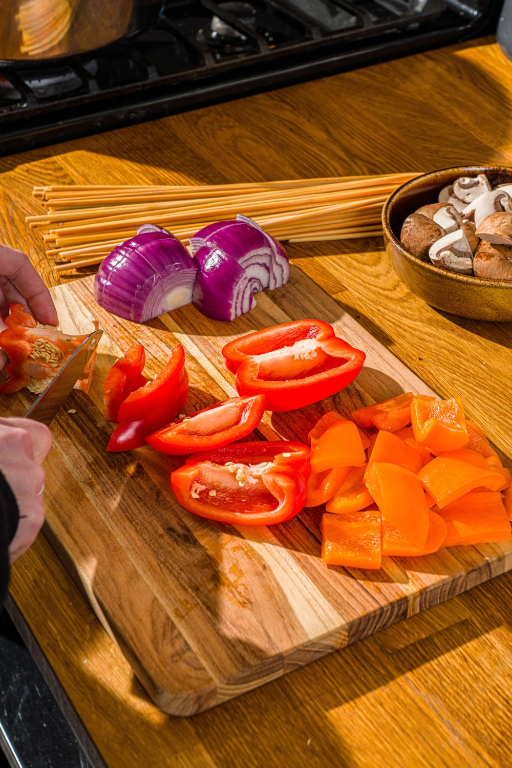 A wooden cutting board with orange and red peppers being chopped. The board is on a wooden counter with a bowl of mushrooms and skewers.