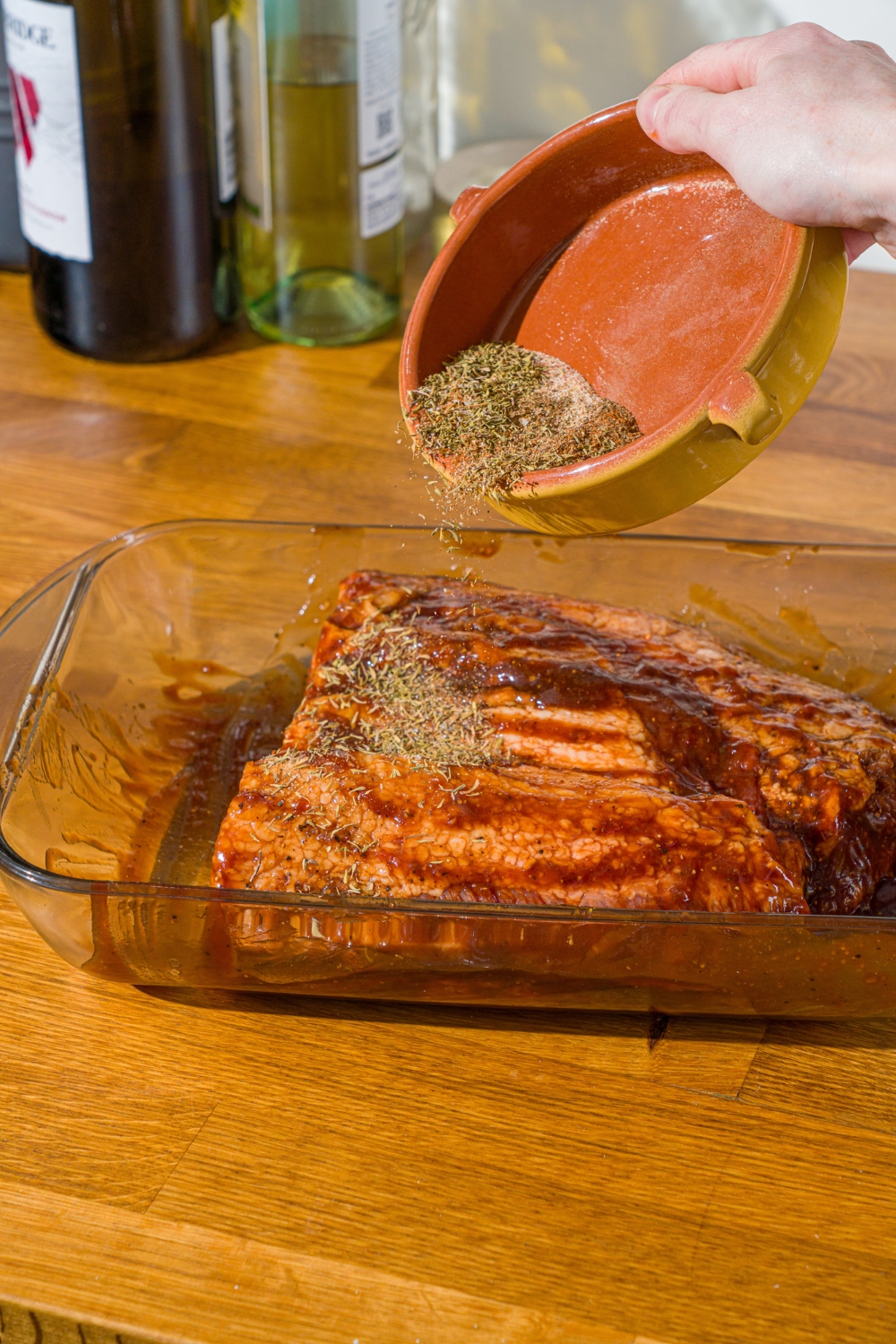 An uncooked beef brisket covered in bbq sauce in a glass baking dish. A bowl of seasonings is being poured over the brisket. The dish is on a wooden counter.