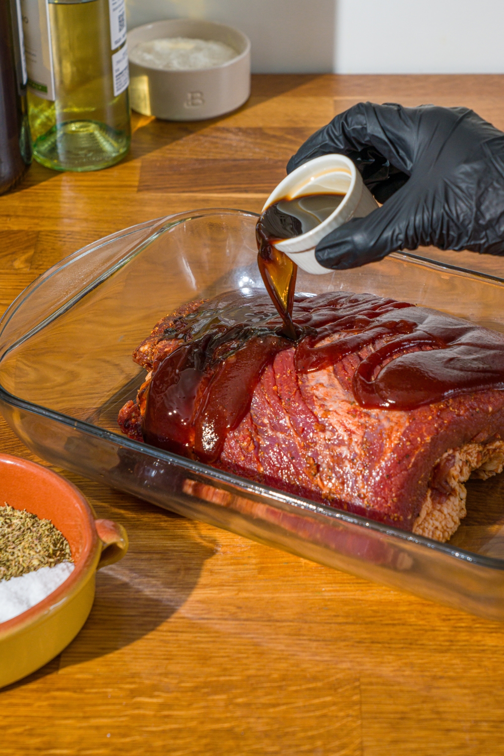 A glass baking dish with an uncooked beef brisket. A bowl of sauce is being poured over the brisket. The dish is on a wooden counter.