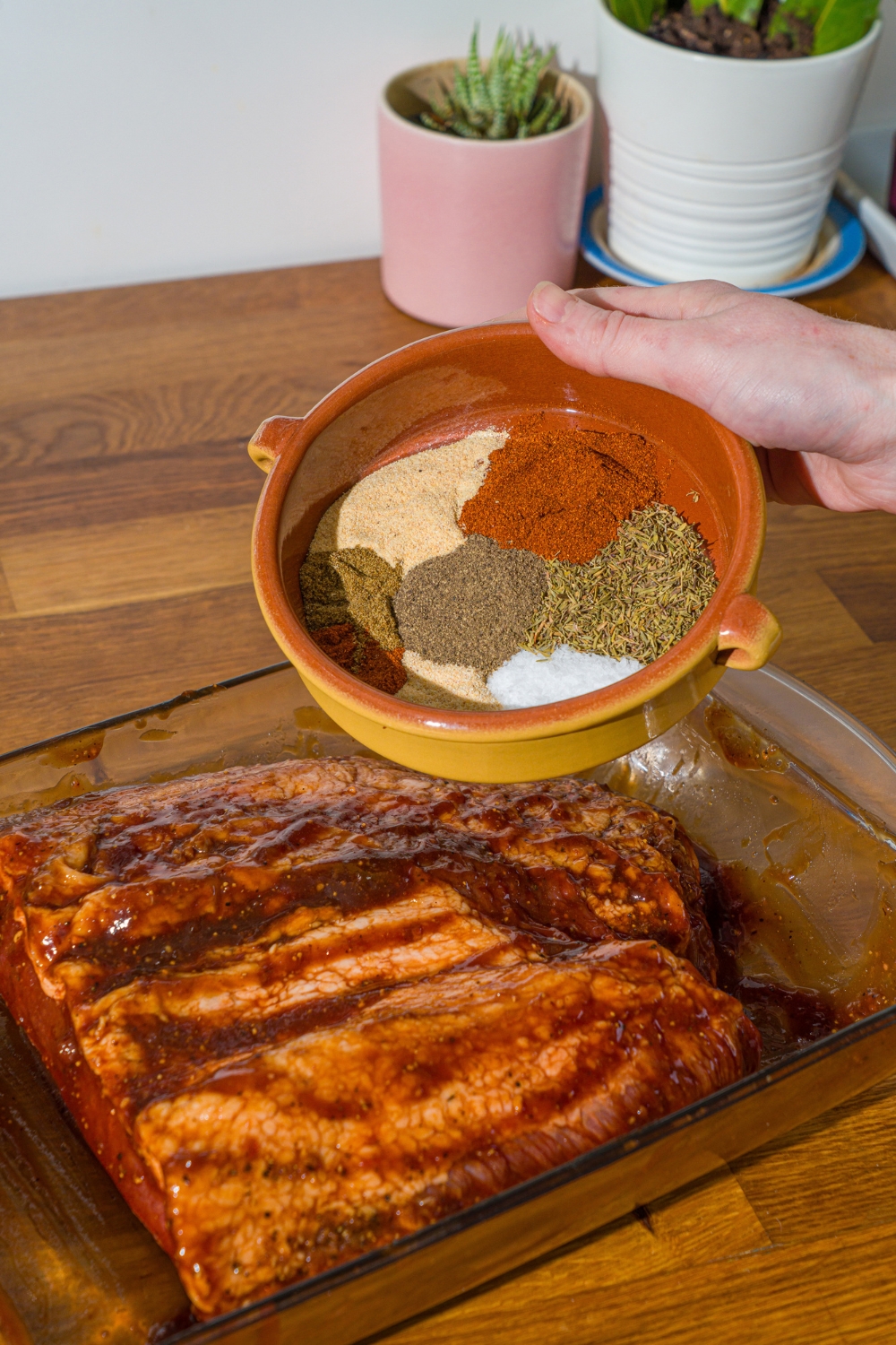 An uncooked beef brisket covered in bbq sauce in a glass baking dish. A bowl of seasonings is held over the dish. The dish is on a wooden counter.