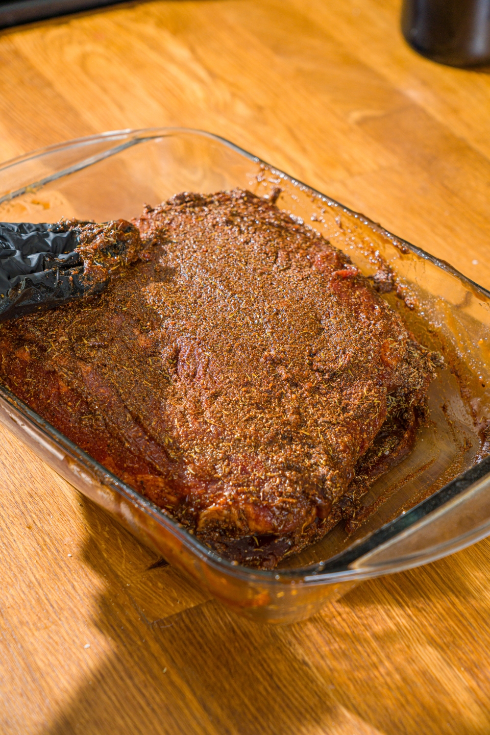 A glass baking dish with uncooked beef brisket topped with bbq sauce. A hand is rubbing seasonings into the brisket.