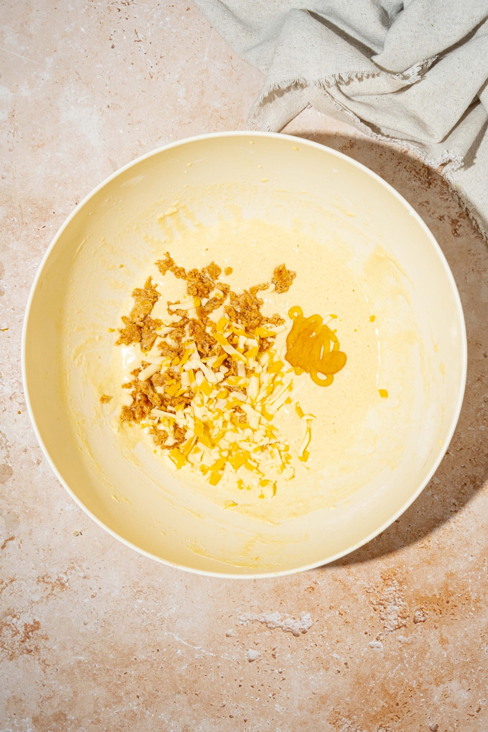 A large white bowl with pancake batter topped with sausage, maple syrup, and shredded cheddar cheese. The bowl is on a tan counter.