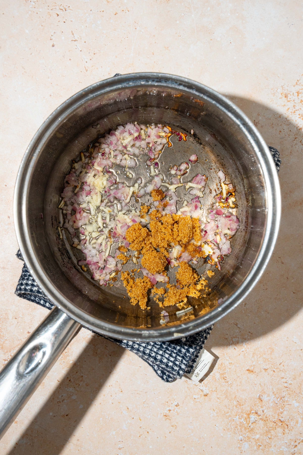 A pot with sautéed shallots, garlic, and brown sugar in oil. The pot is on a tan counter with a dish towel.