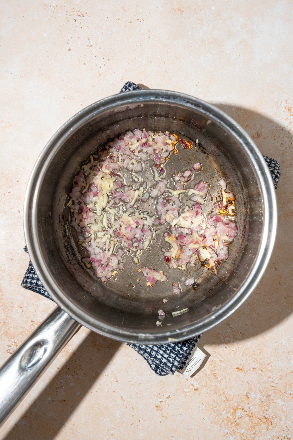A pot with shallots and garlic sautéing in oil. The pot is on a tan counter with a dish towel.