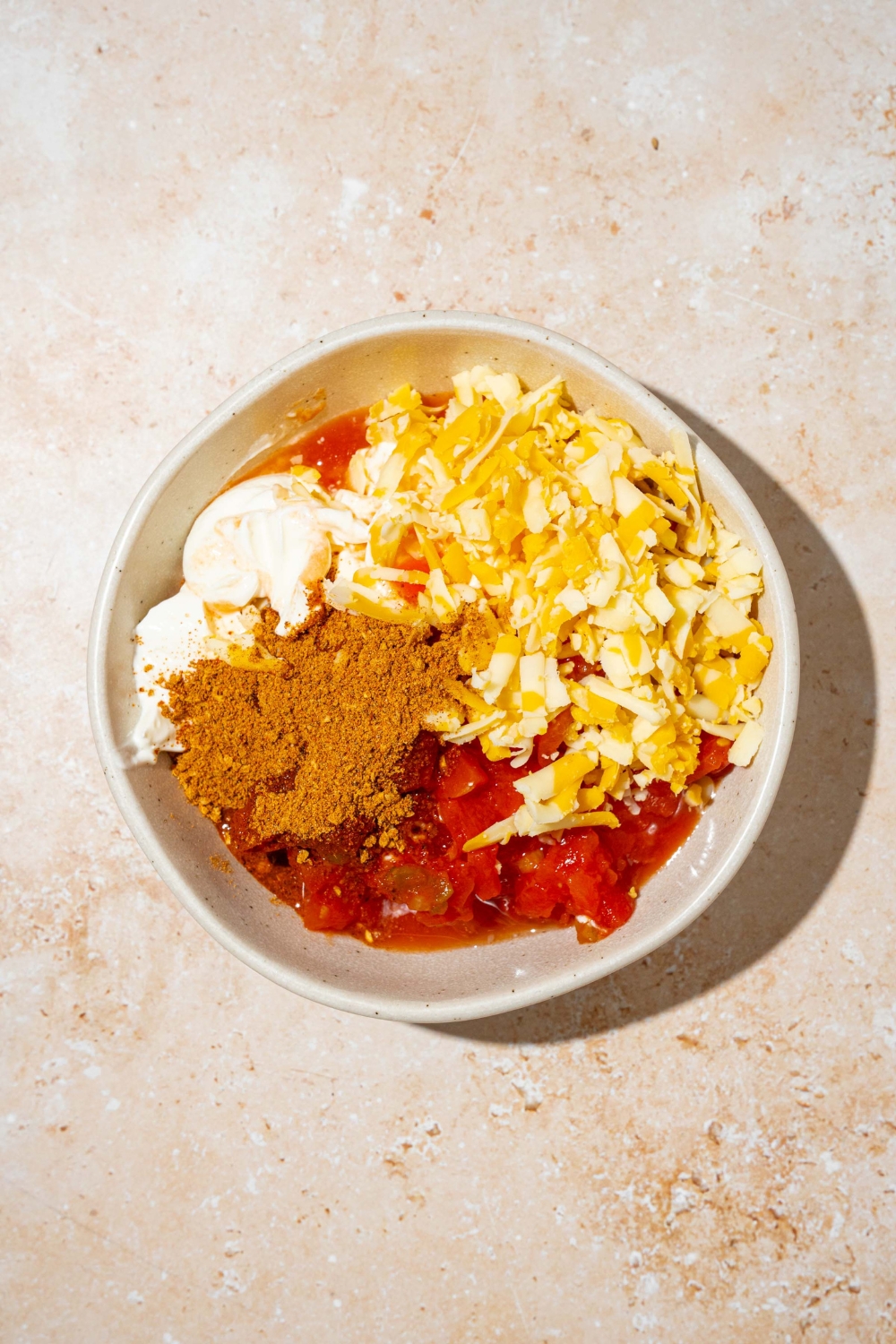 A white bowl with ingredients to make poolside dip including sour cream, rotel, taco seasoning, and shredded cheese. The bowl is on a tan counter.