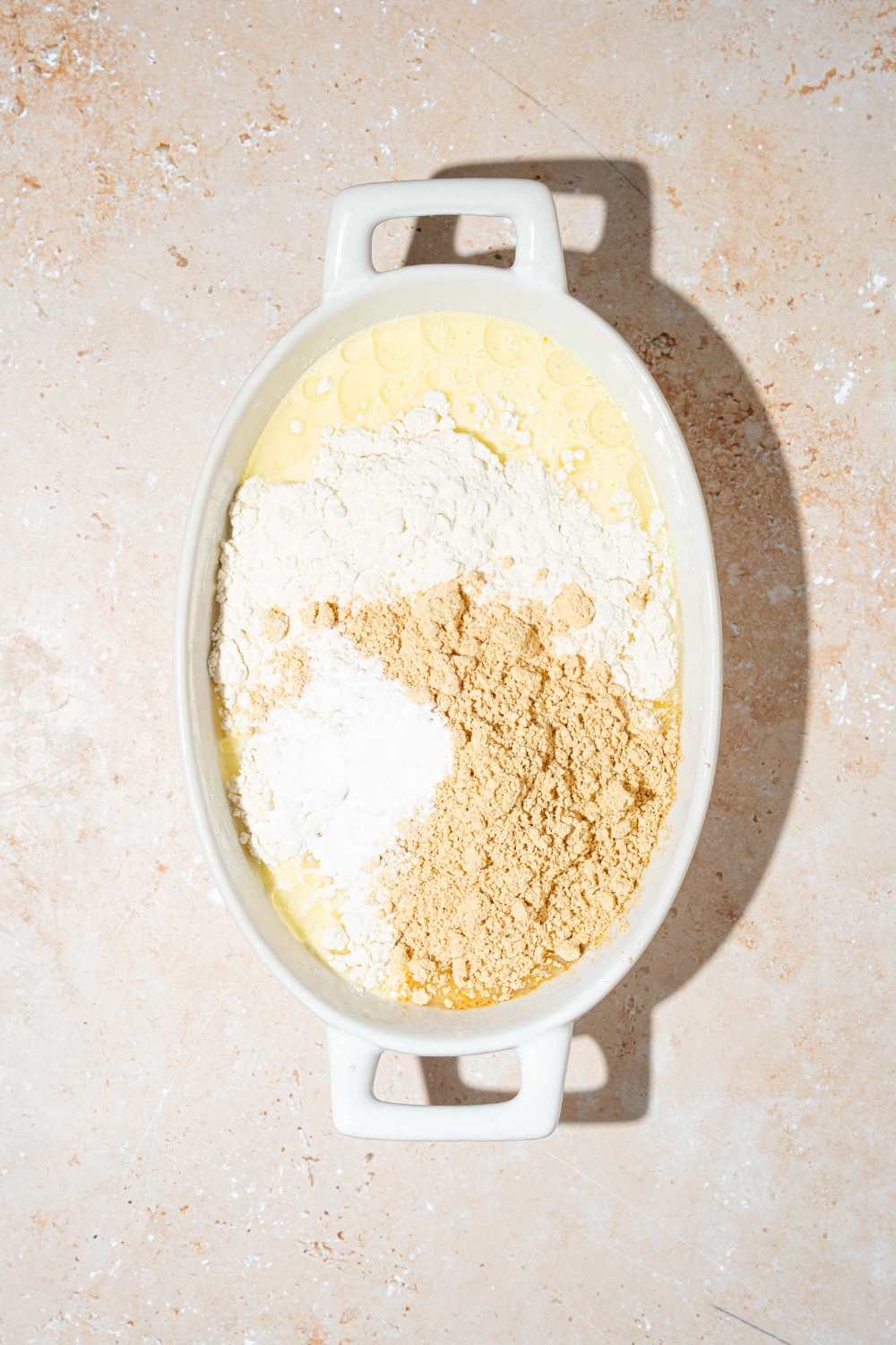 An oven safe bowl with dry ingredients for protein pancake batter over a wet ingredient mixture. The bowl is on a tan counter.