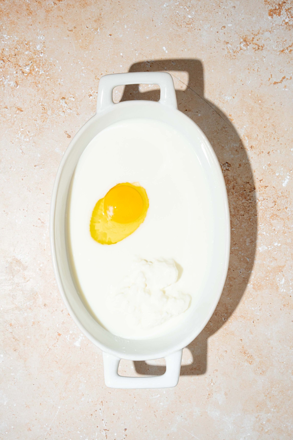 An oven safe bowl with wet ingredients to make protein pancake batter like egg, yogurt, and milk. The bowl is on a tan counter.