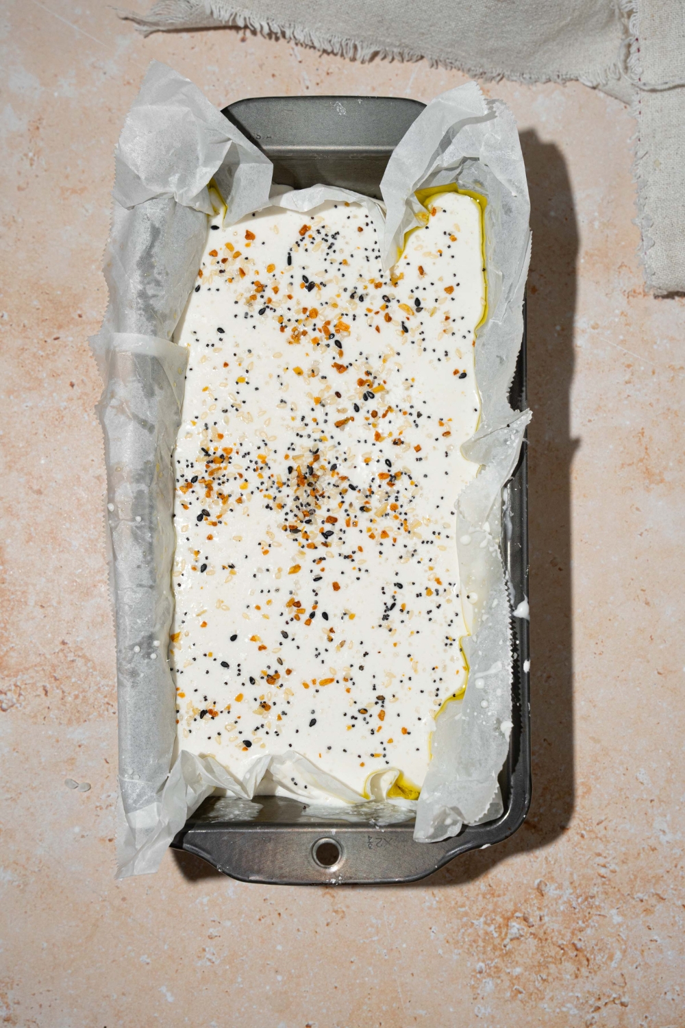 A loaf pan lined with parchment paper with rice blender bread dough topped with everything bagel seeds. The pan is on a tan counter.